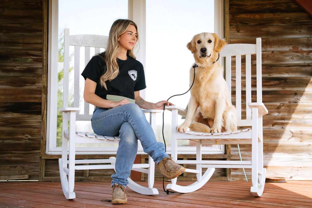 A trainer and a golden retriever sit in white rocking chairs on a rustic wood porch